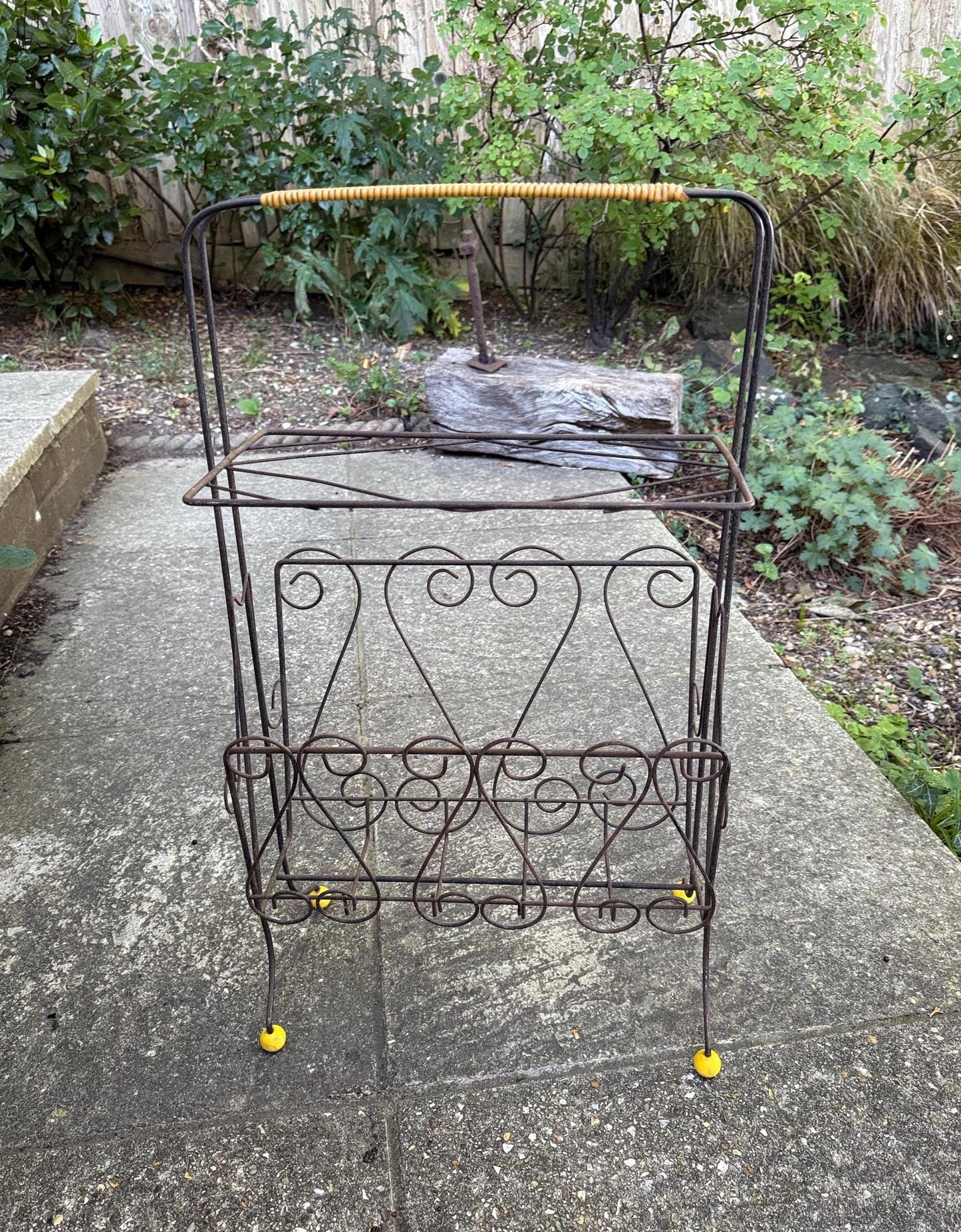 a mid century scrollwork black metal work storage rack. with a plastic yellow wrap around handle and yellow round feet. Very retro and mid century. Rusted finish currently
